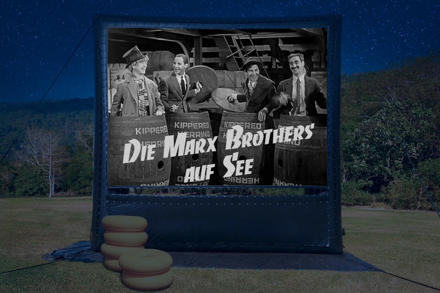 Das Bild zeigt die Nachtaufnahme einer aufblasbaren Videoleinwand auf einer grünen Wiese. Die Leinwand zeigt ein Standbild aus dem Film 'Die Marx Brothers auf See', das die vier Marx Brothers in Holzfässern stehend zeigt und den Schriftzug 'Die Marx Brothers auf See'.