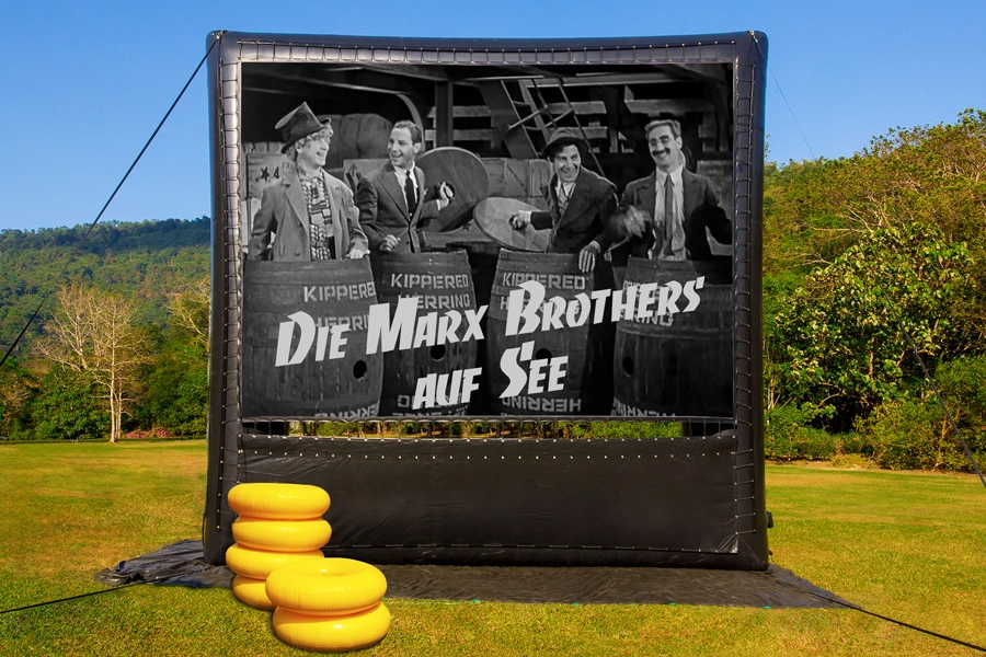Das Bild zeigt eine aufblasbare Videoleinwand auf einer grünen Wiese. Die Leinwand zeigt ein Standbild aus dem Film 'Die Marx Brothers auf See', das die vier Marx Brothers in Holzfässern stehend zeigt und den Schriftzug 'Die Marx Brothers auf See'.